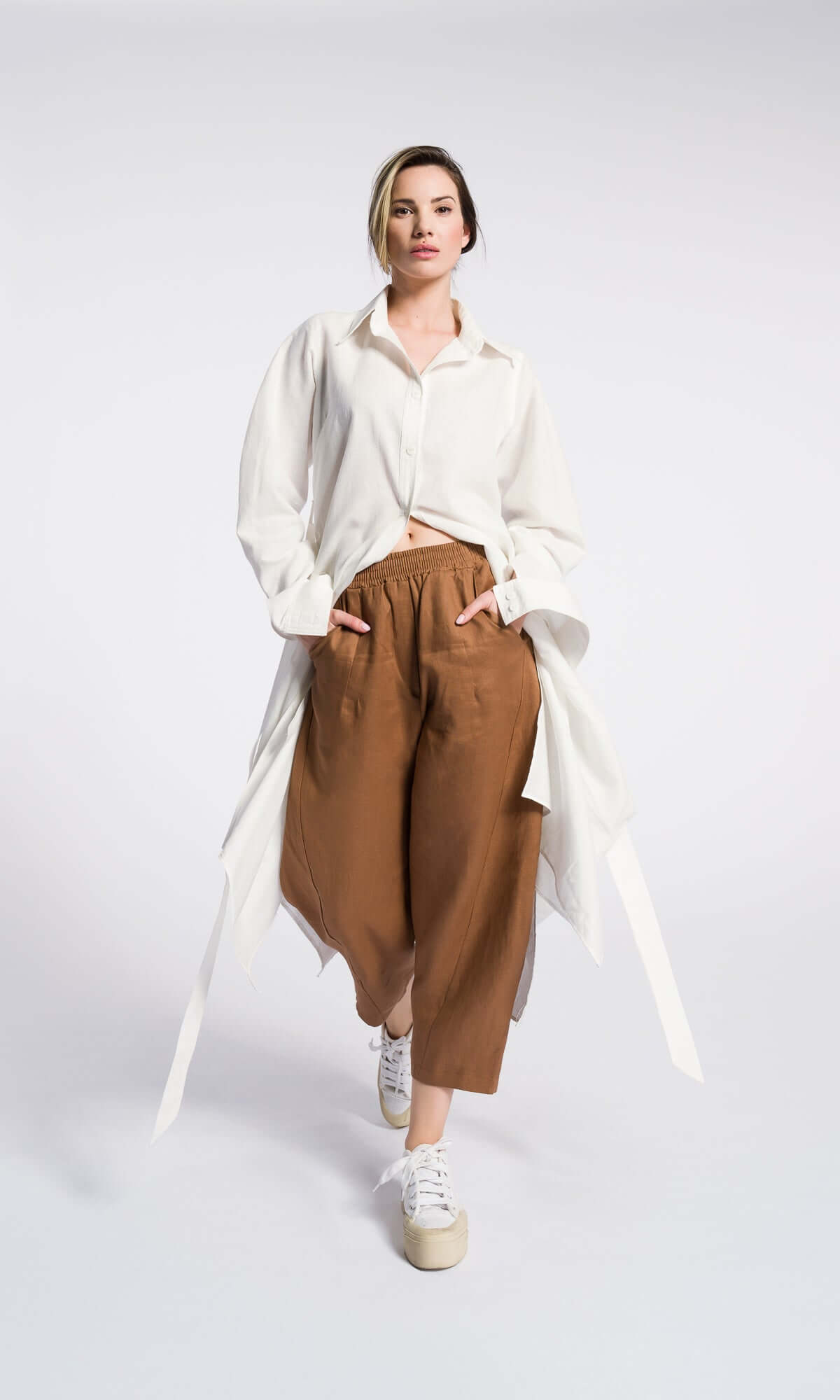 A model wearing curved leg tobacco color linen pants with a cropped length and elastic waistband, side pockets paired with a white shirt and white sneakers.