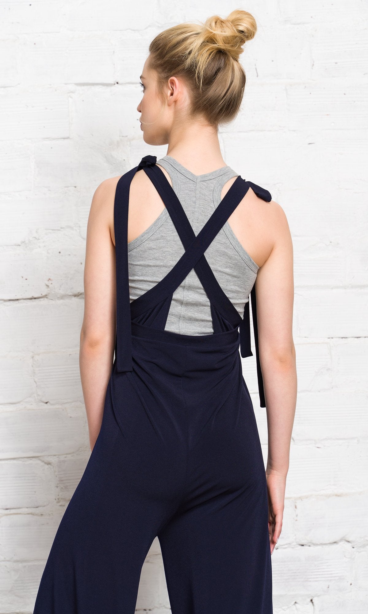 Back view of women's navy blue wide-leg jumpsuit overalls featuring criss-cross back straps over a grey casual tank top.