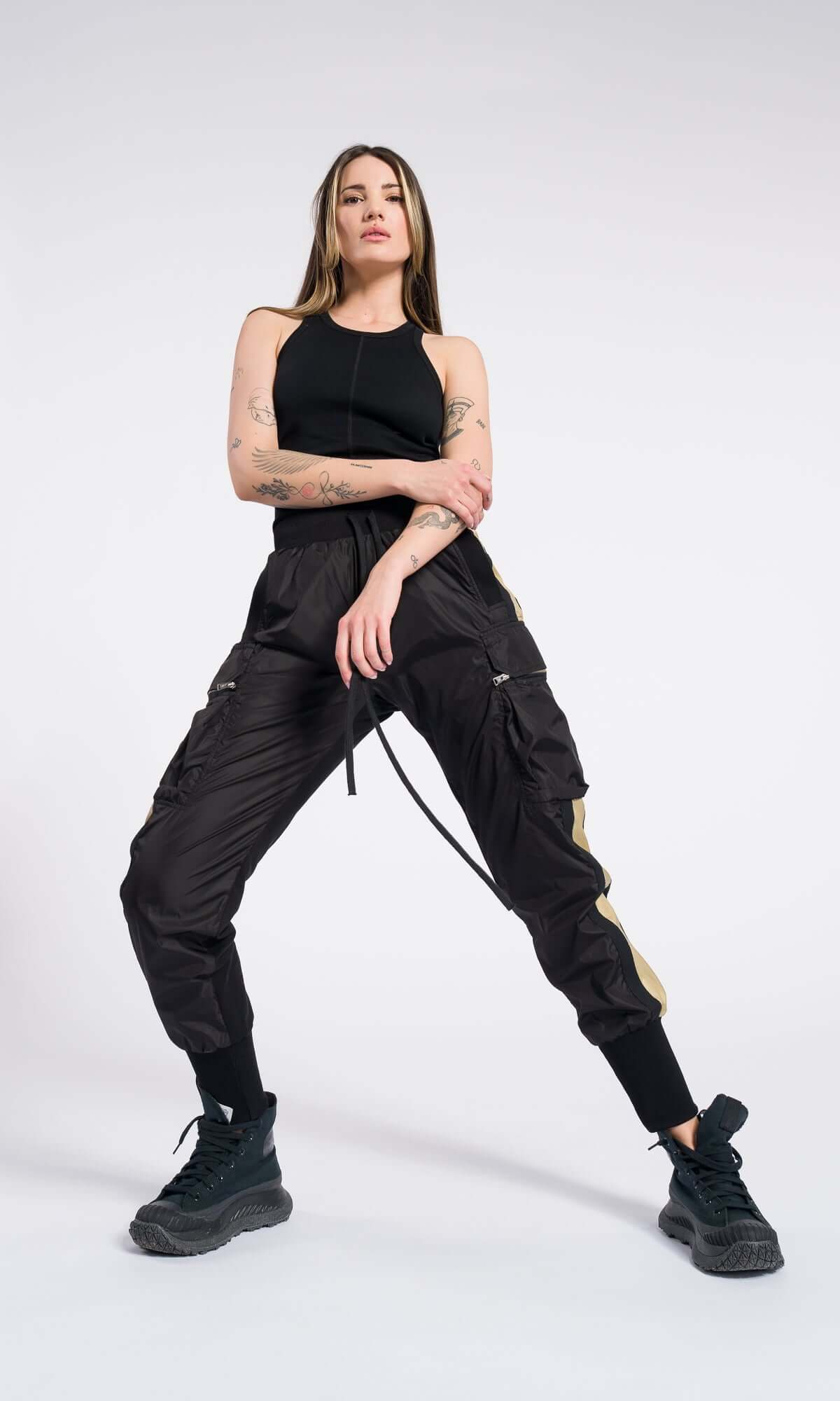 woman posing with a wide stance, wearing a black top, combat boots, and striped cargo pants