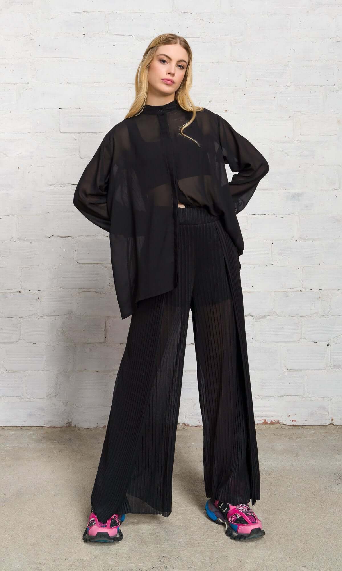 Front view of a blonde model standing in a black sheer button-up shirt and wide-leg trousers