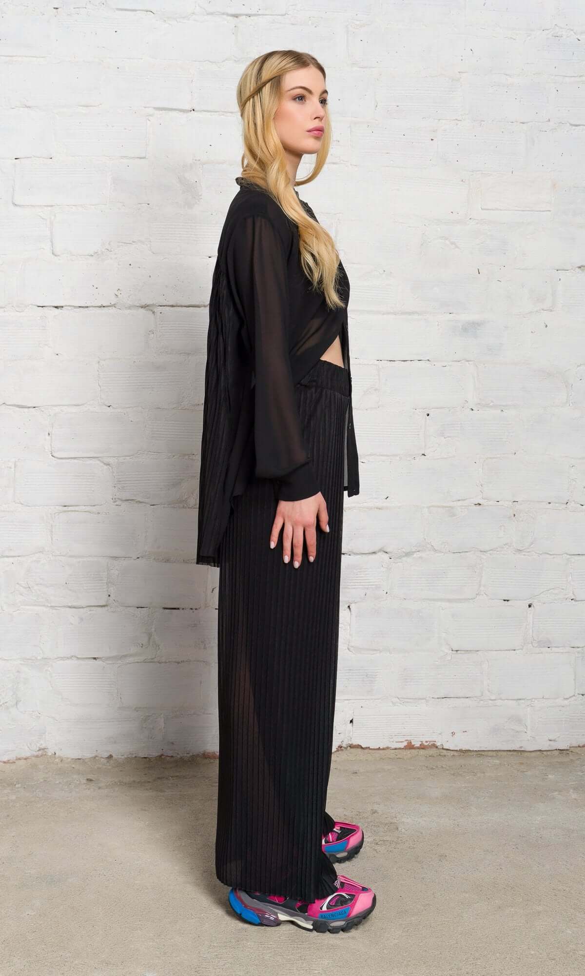 Side profile of a blonde model standing against a white brick wall in a black sheer outfit