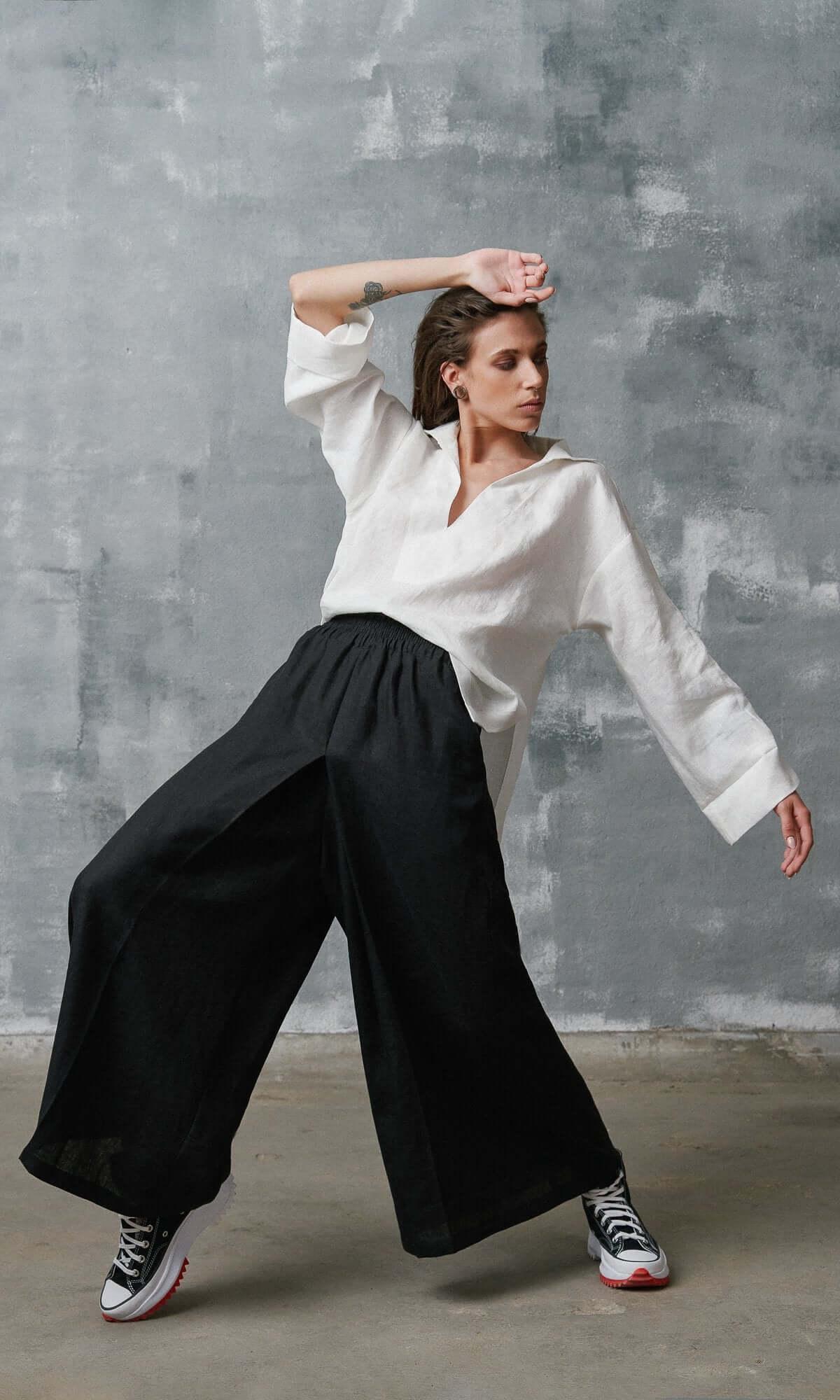 A model wearing a white shirt and black linen skirt pants with an elastic waistband and inverted pleats, standing against a plain background, showing wide leg loose fit .