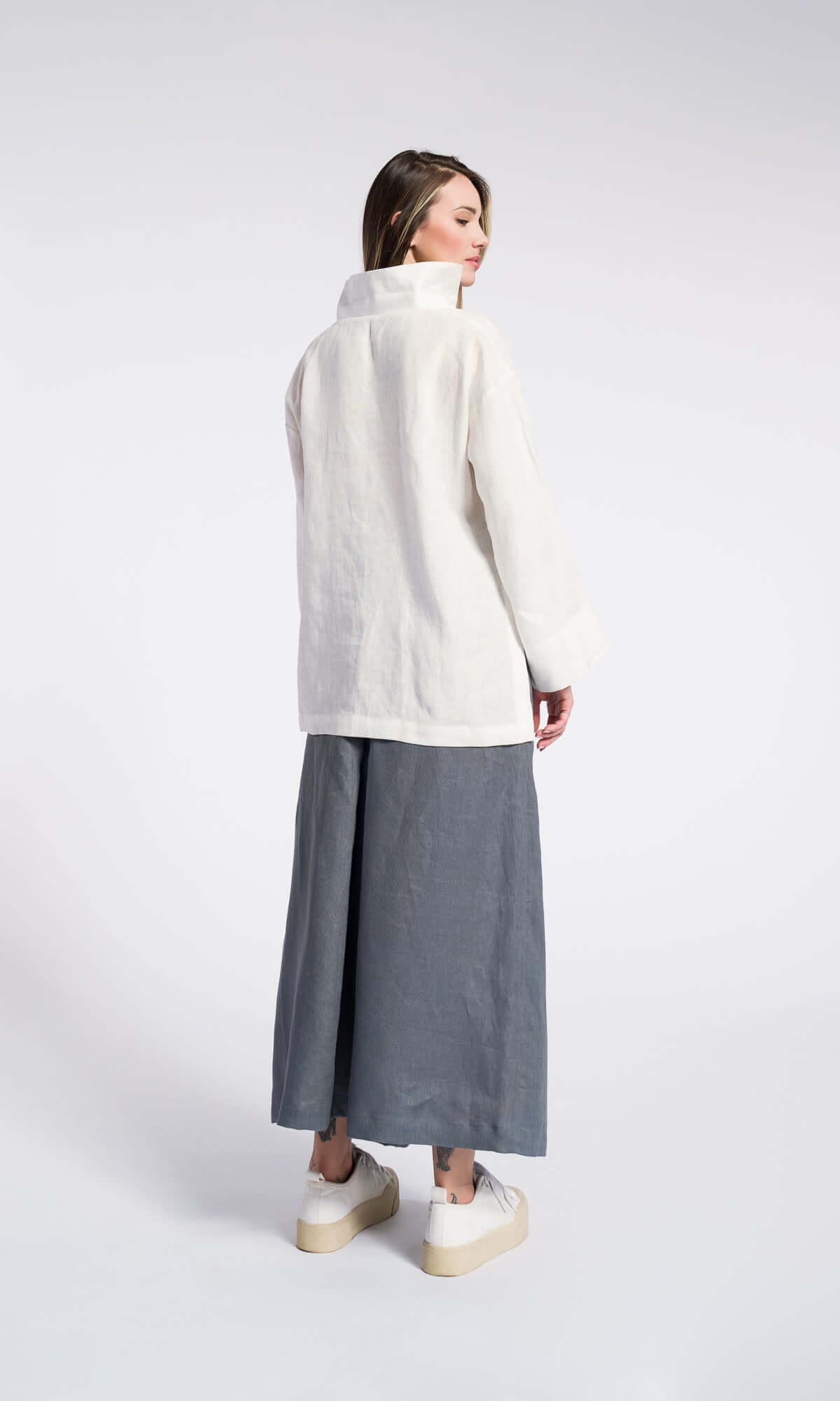 Back side view of a model wearing a white shirt and grey linen skirt pants with an elastic waistband and inverted pleats, standing towards a plain background.
