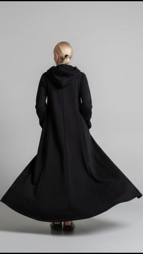 Person wearing a long black dress standing in a minimalistic room with a white wall and gray floor.