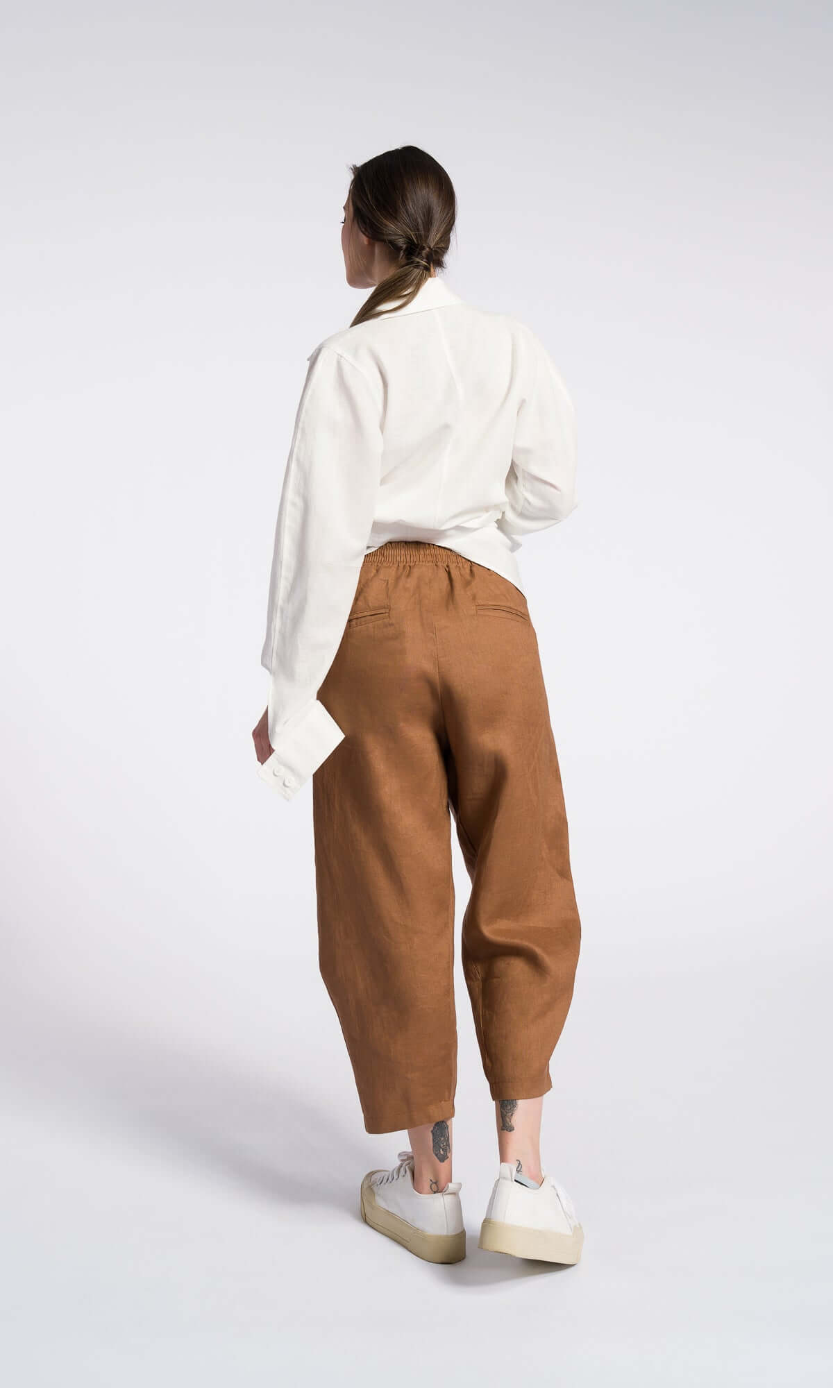 Back side view of a model wearing curved leg linen pants with a cropped length and elastic waistband, back pockets paired with a white shirt and white sneakers.
