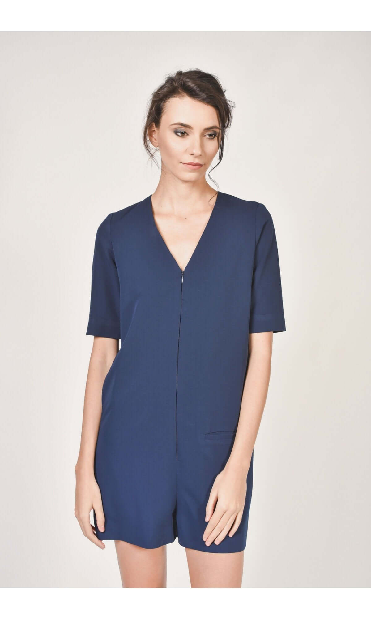 A woman wearing a navy blue short jumpsuit with a V-neckline, short sleeves, and front pockets, zipper front, standing against a neutral background.