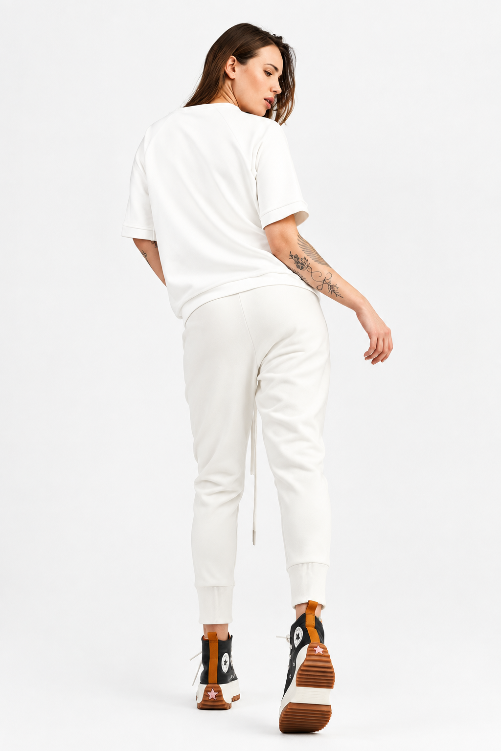 A woman is shown from behind wearing a white t-shirt with a black triangle graphic on the back, black jogger pants, and black and orange sneakers, set against a plain light gray studio background.