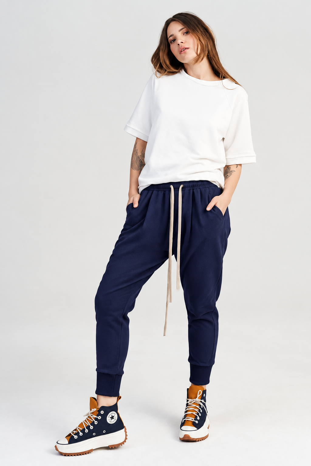 A woman modeling a white oversized crewneck sweatshirt, black jogger pants with a beige drawstring, and black high-top sneakers with orange laces, set against a plain light gray studio background.