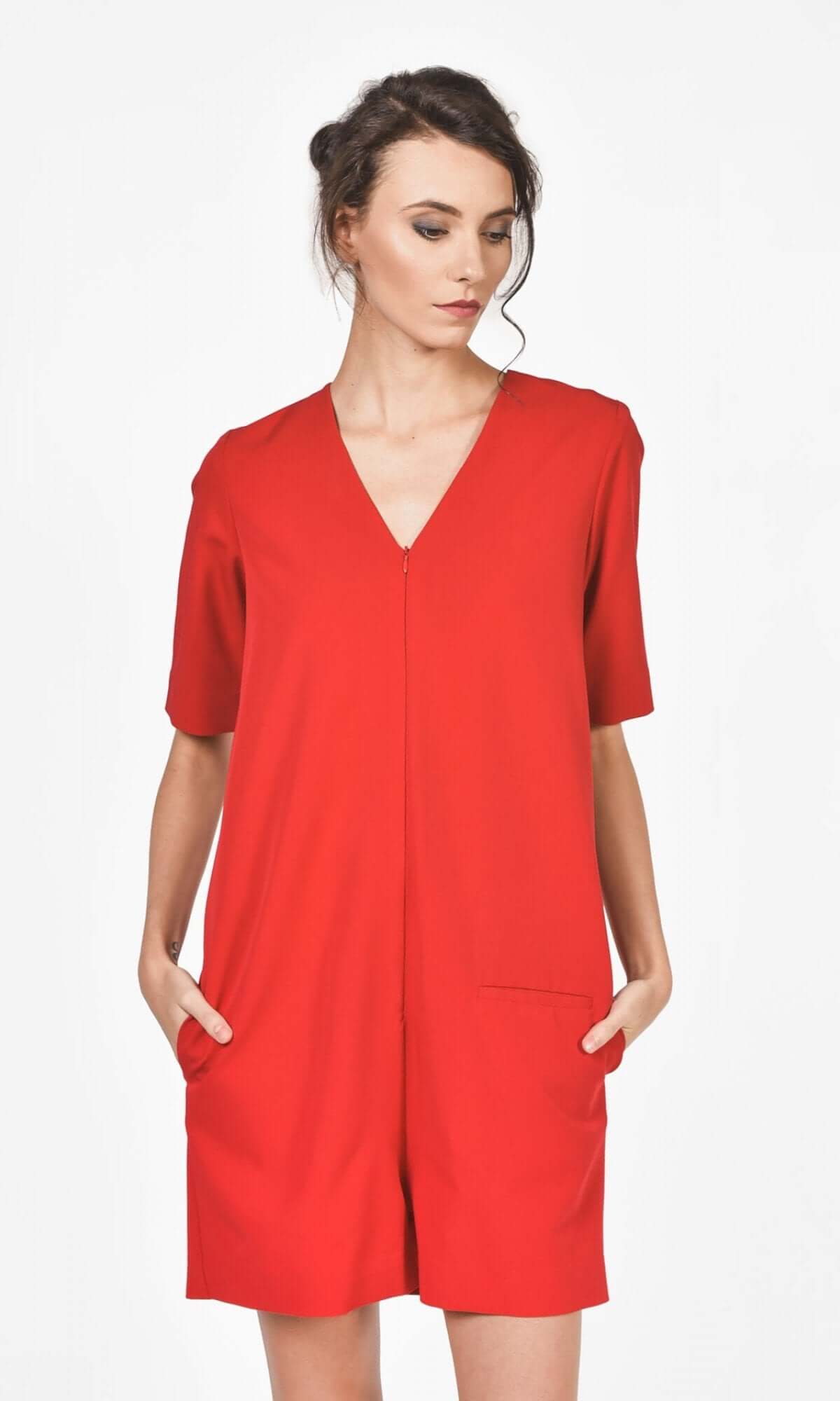 A woman wearing a red short jumpsuit with a V-neckline, short sleeves, and front pockets, zipper front, standing against a neutral background.