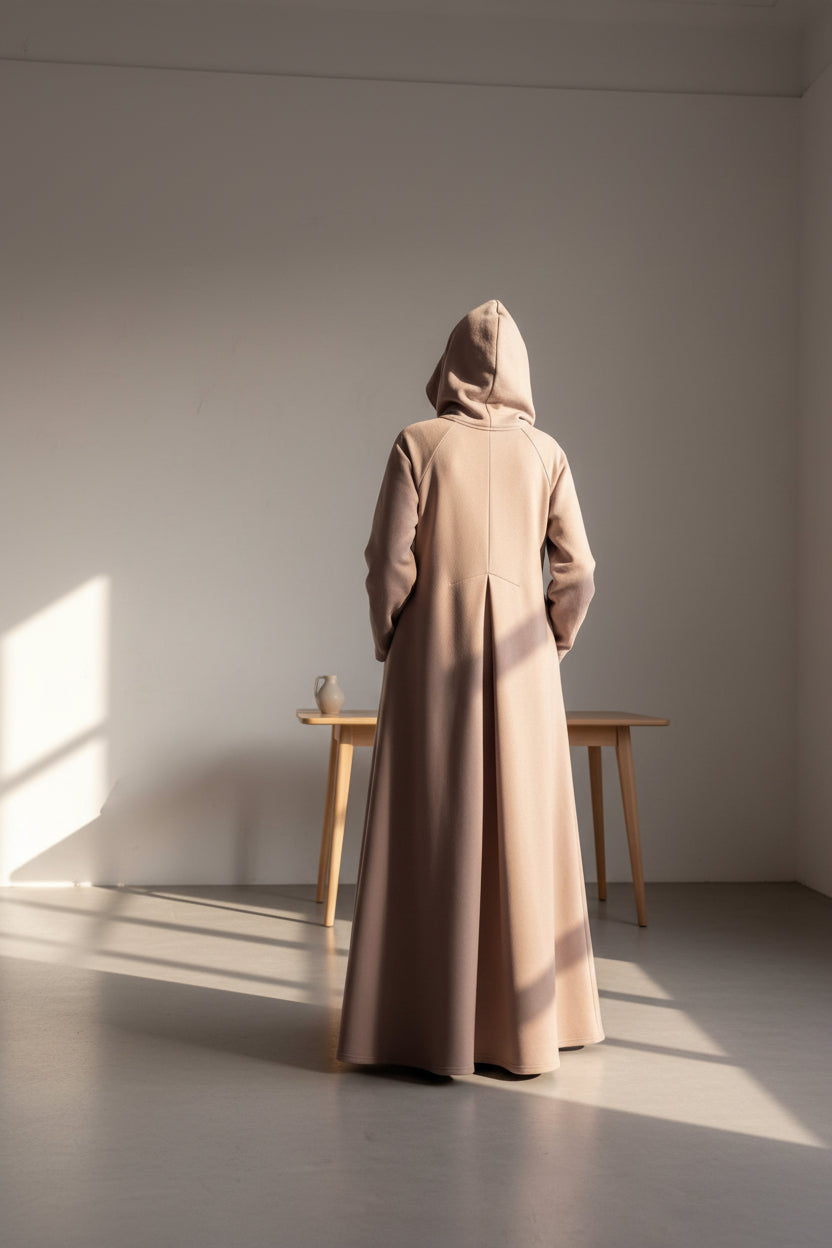 Person wearing a long beige coat standing in a minimalistic room with white walls and floor.