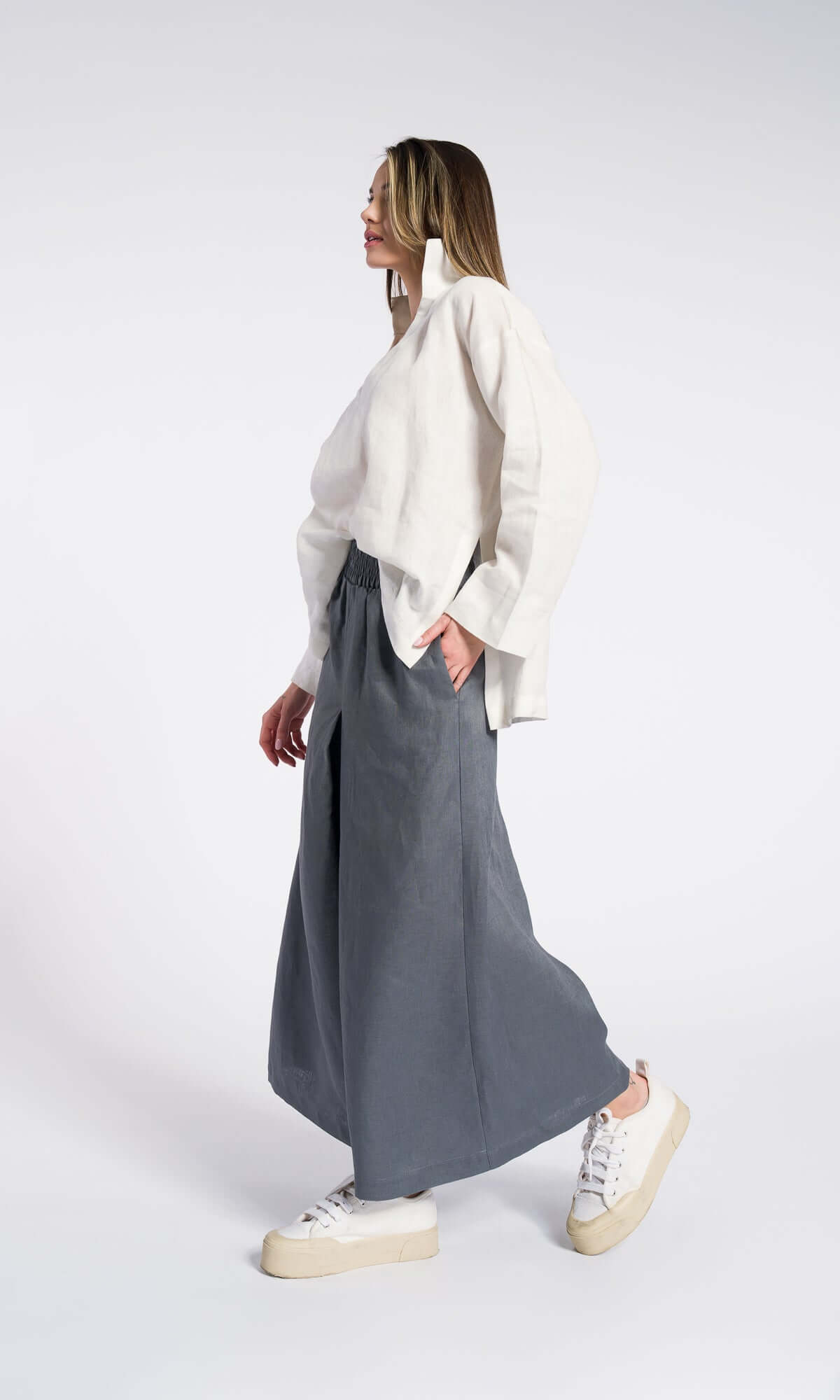 Side view of a model wearing a white shirt and grey linen skirt pants with an elastic waistband and inverted pleats, side pocket and showing loose wide leg and relaxed fit