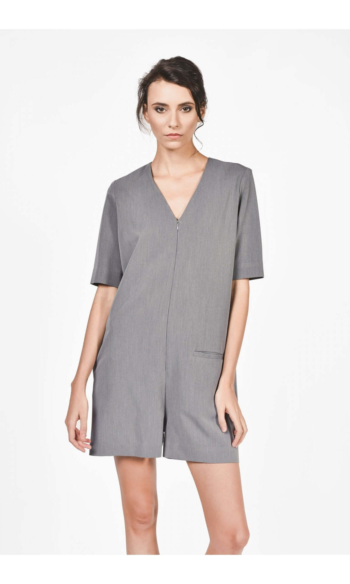 A woman wearing a grey short jumpsuit with a V-neckline, short sleeves, and front pockets, zipper front, standing against a neutral background.