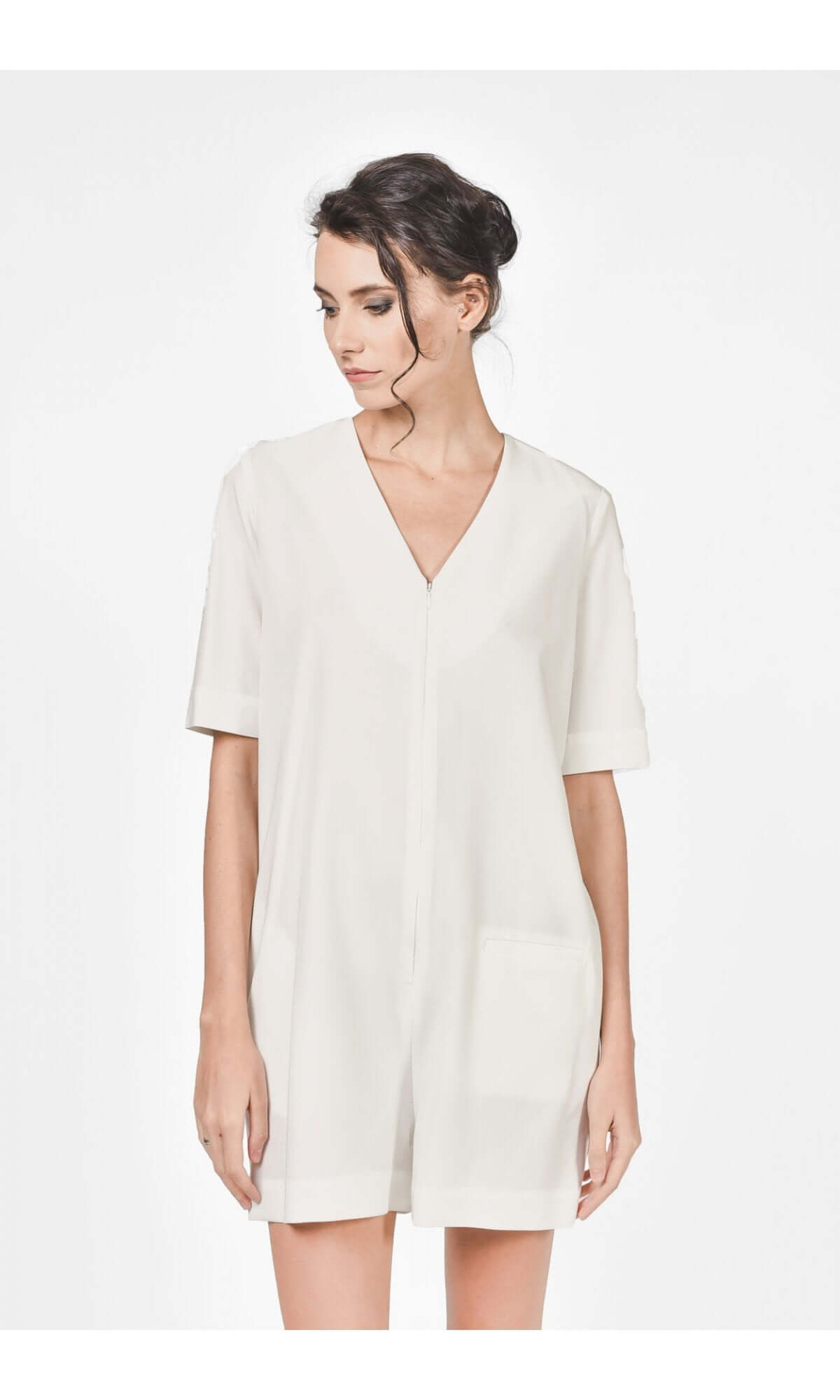 A woman wearing an off white short jumpsuit with a V-neckline, short sleeves, and front pockets, zipper front, standing against a neutral background.