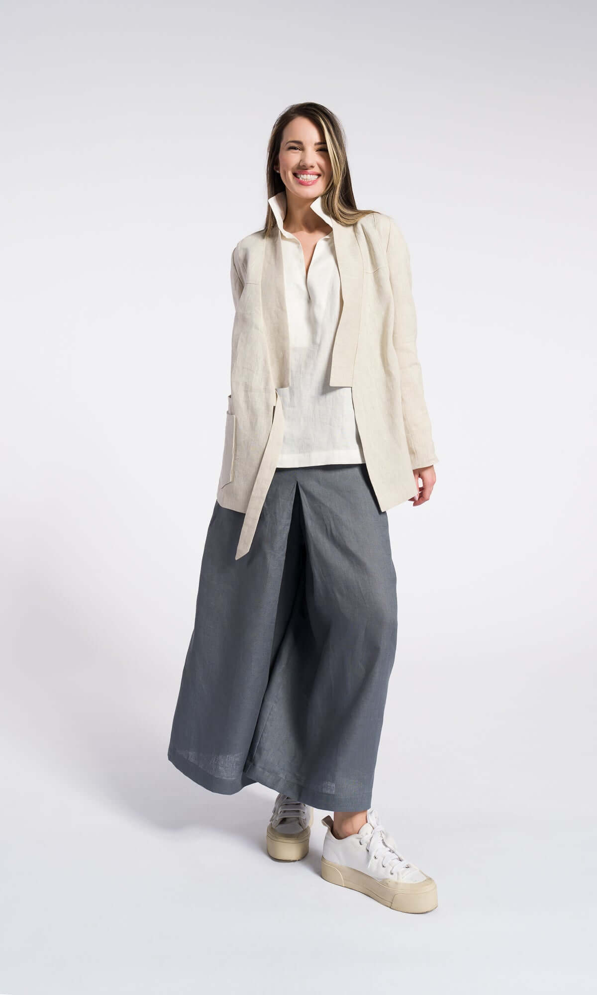 A model wearing a white shirt off white linen blazer over and grey linen skirt pants with an elastic waistband and inverted pleats, standing against a plain background. Loose relaxed fit showing