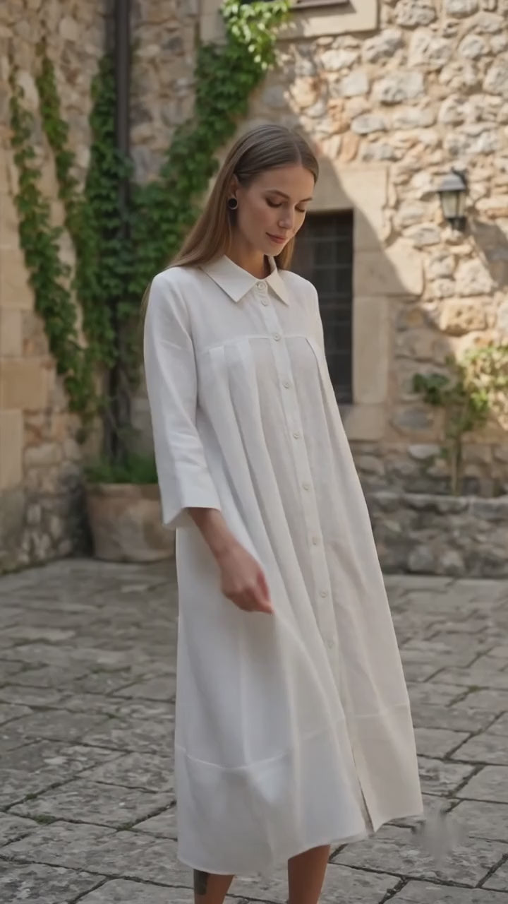 Woman looking down while posing in a flowing, long-sleeved white shirt dress outdoors